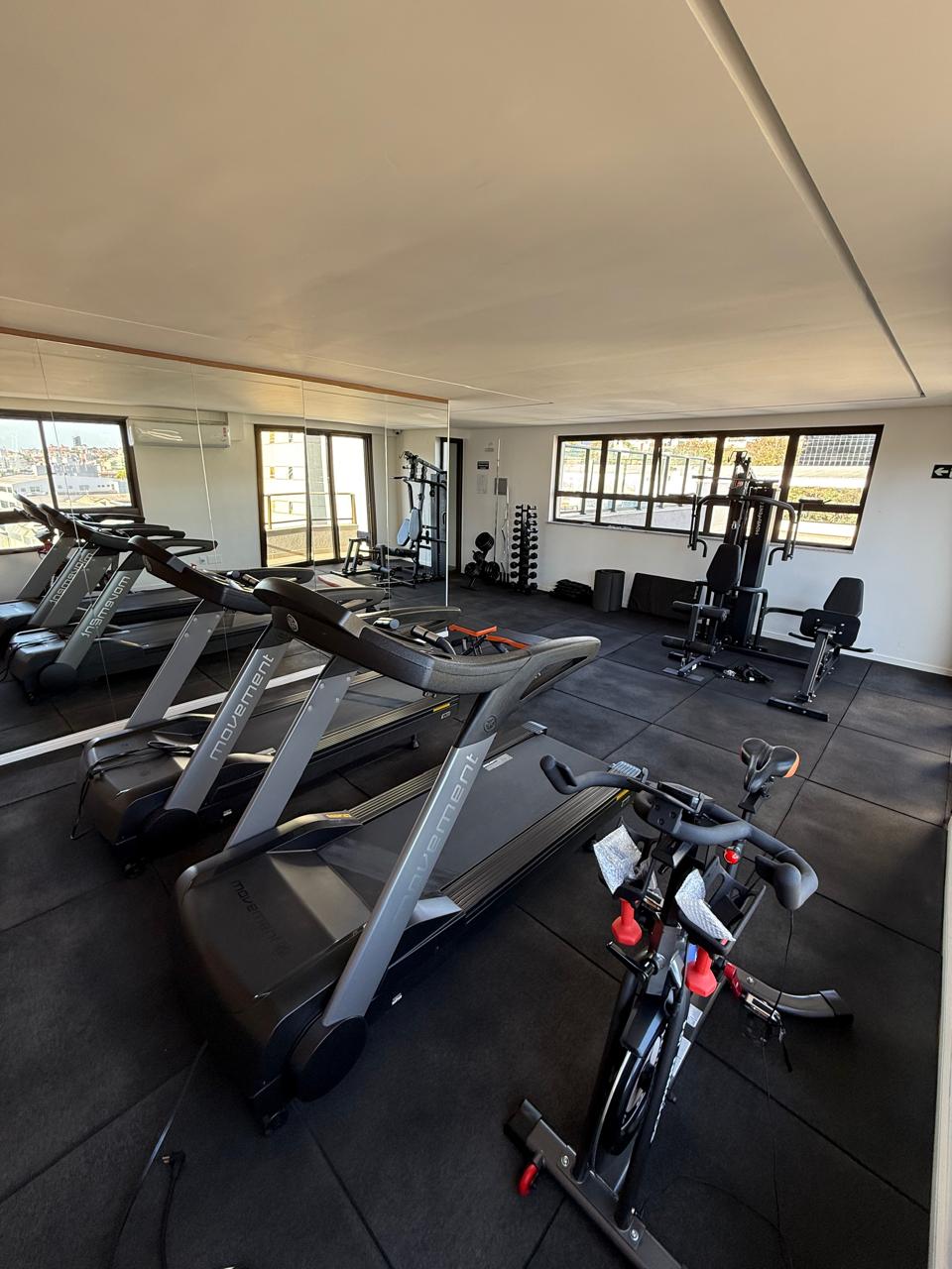 Espaço fitness com esteiras, pesos livres e amplas janelas